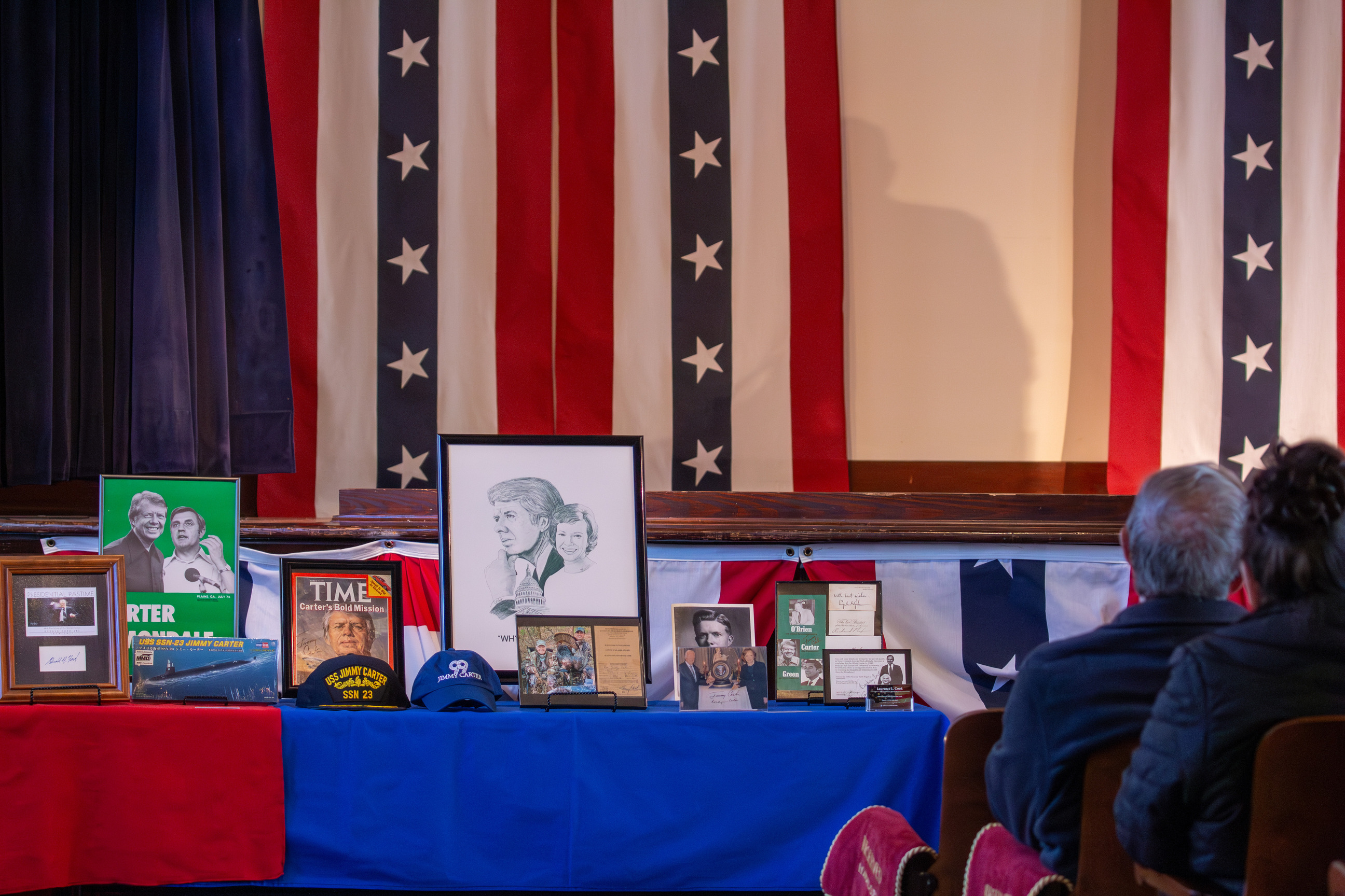 Presidential memorabilia on table. People sitting in auditorium seats on right. Red, white, and blue flag banners in background.