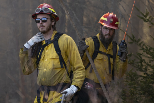 Two firefighters in protective gear with red helmets, surrounded by smoke, with one using a radio. They are standing in a forested area with smoke in the air.
