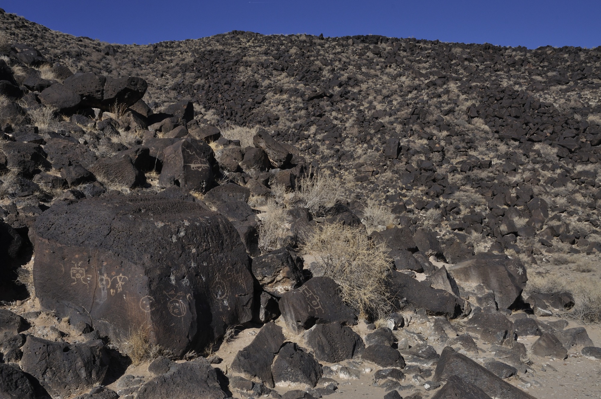 Photograph of blocks of large basalt blocks with petroglyphs carved into the dark patina of desert varnish on the large boulders, exposing the lighter colored basaltic rock beneath. 