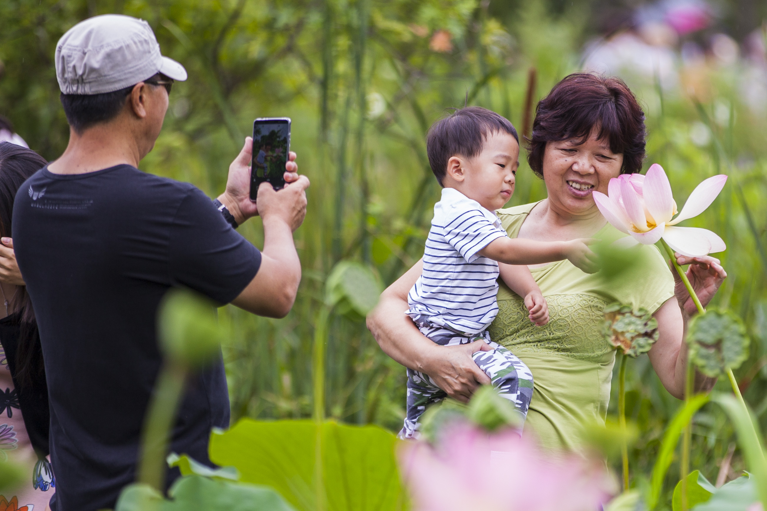 A man takes a photo of an older woman with a young child touching a lotus flower