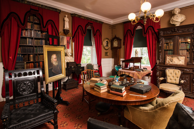 View of a study with a round center table cluttered with books and writing implements. A standing desk sits on a second round table in front of a window. The room has brown wallpaper and deep red curtains and carpet. An ornately carved black armchair is in the foreground.