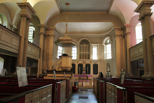 An interior image of King's Chapel, the space is slightly dark. The photographer is situated in the middle aisle of the church's first floor. There are multiple box pews along the aisle going forward to the pulpit, which is raised above the floor. Above the pulpit is a sounding board to help voices carry. Behind the pulpit are busts, candles, and other liturgical items. The ceiling is vaulted and there are small portions of the 2nd level balcony included in the image.