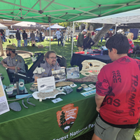Rangers at a booth with green canopy and tables with various items help people, many other booths in background.