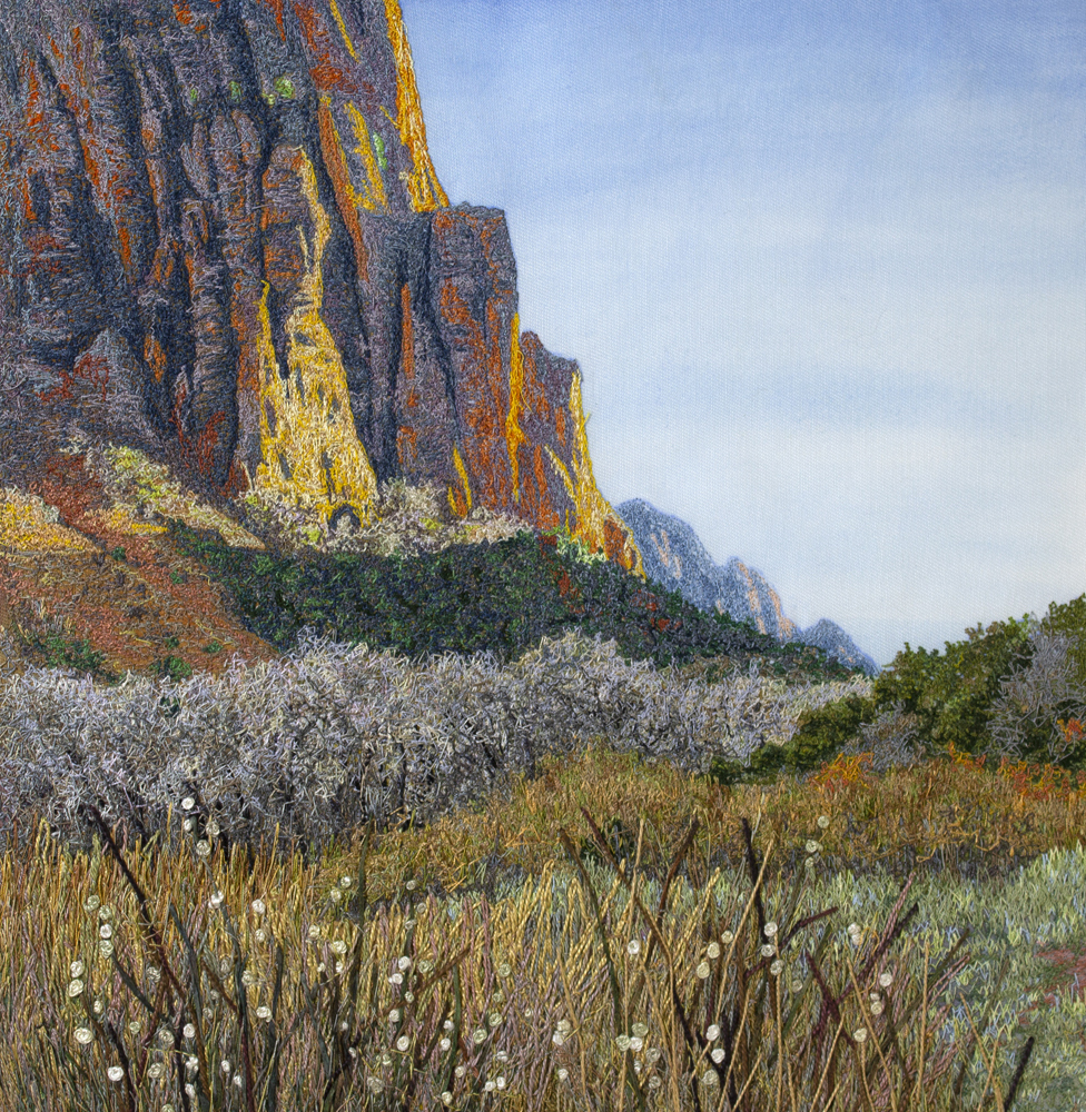 An image created using colored thread and fabric. A field of grasses, bushes, and shrubs gives way to a hillside covered in greenery. Small strips of loose sediment scar the hillside. Above the hillside, a tall sandstone cliff reaches out of frame. In the far background, other sandstone peaks are visible beneath a blue sky.