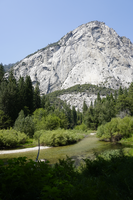 A slow flowing river cuts through the lush plants and trees that surround the river. Behind the river rises high into the sky , a large granite mountain. 