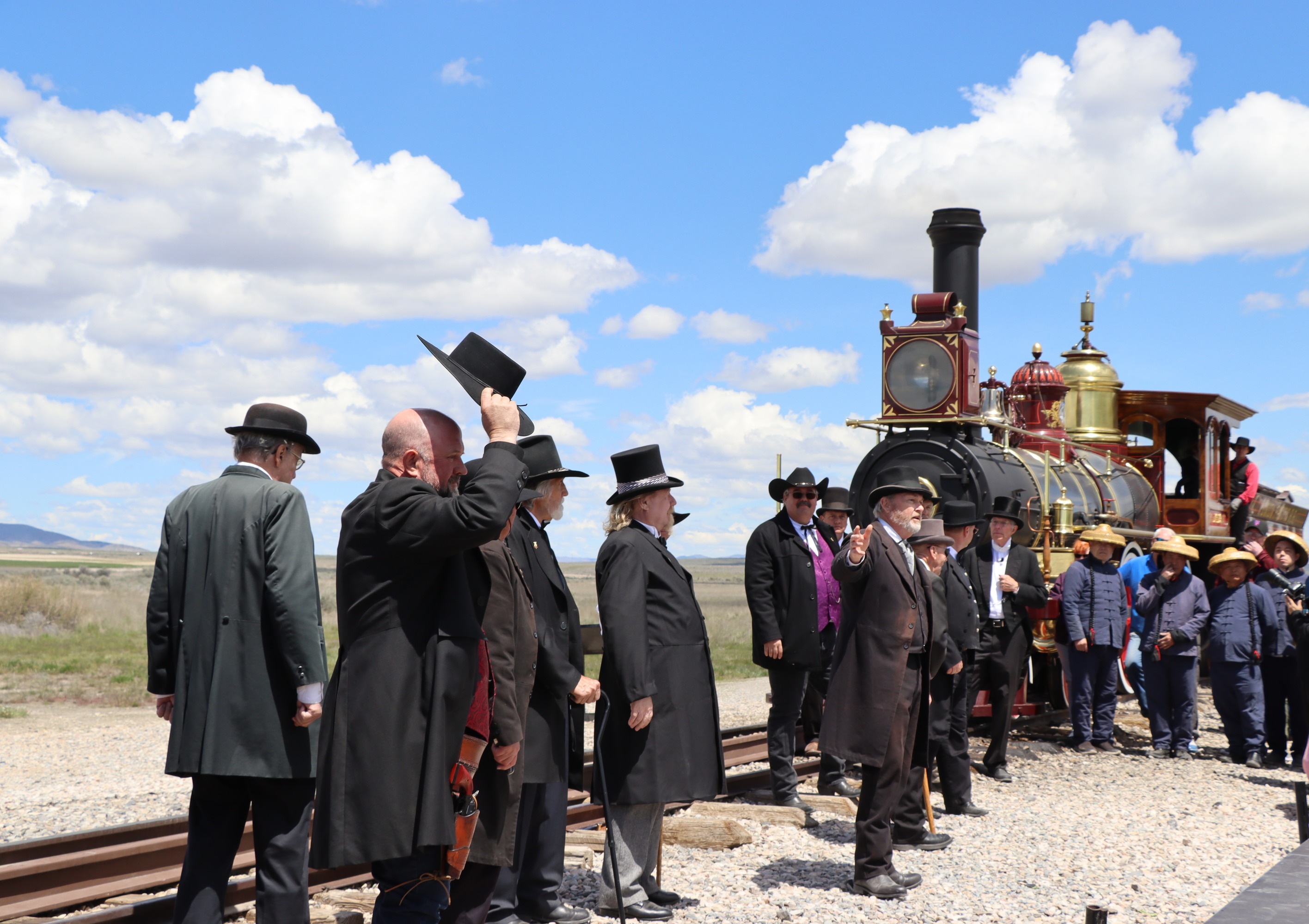 Living history volunteers in 1860's period dress take hats off as one addresses the crowd. No. 119 a red Victorian steam locomotive sits directly behind living history volunteers. Blue skies can be seen in background. 
