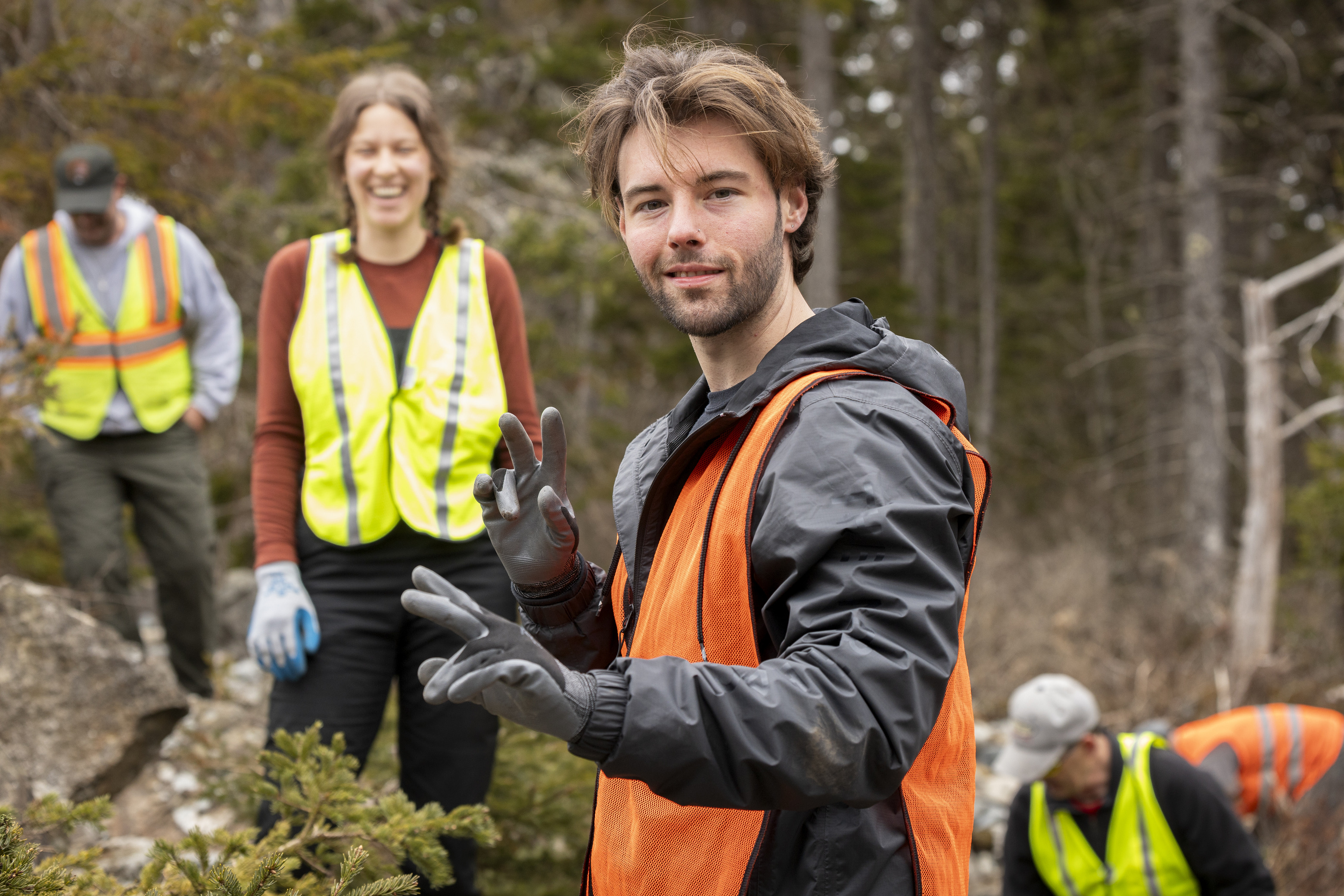Volunteers in reflective vests working in a wooded area of Acadia National Park, with a college-aged male volunteer in the foreground making a peace sign to the camera.