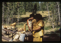 Two women stand at a table holding woven baskets in front of a meadow. One woman holds a basket with a doll to show a child carrier.