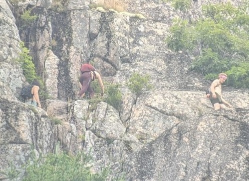 Three suspects partway up the Precipice Trail. One suspect presents as male, and he has chin length wavy hair and facial hair. A person who presents as female wearing purple leggings is just in front of him. A third individual who presents as male with short hair leads the pack across the cliff face.