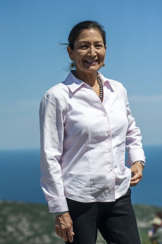 Woman wearing pale pink blouse smiles wide with blue sky and vast ocean behind her.