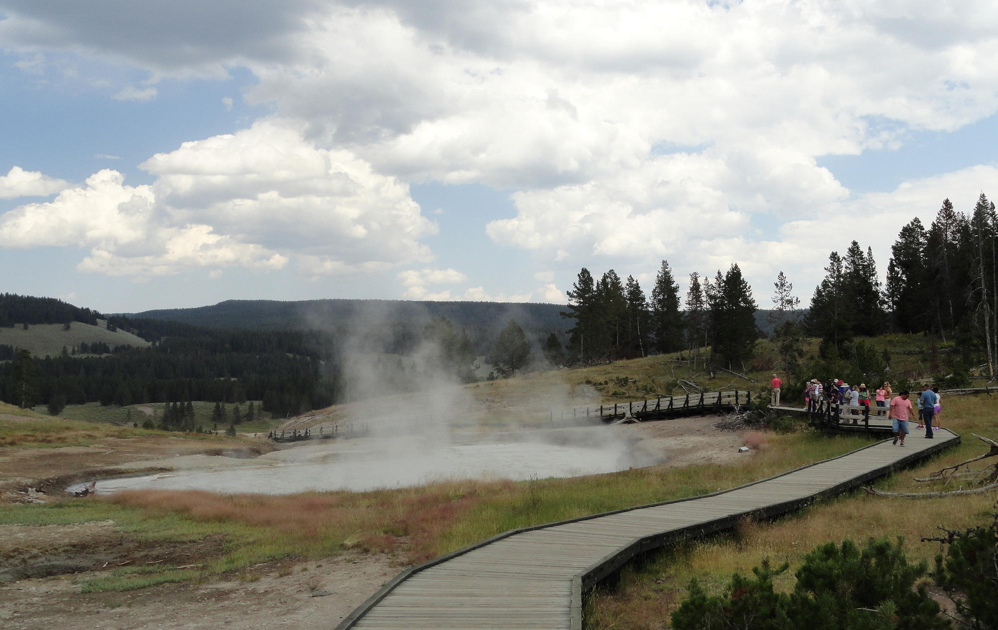 People stand and walk on the boardwalk next to a muddy, steaming hot spring.