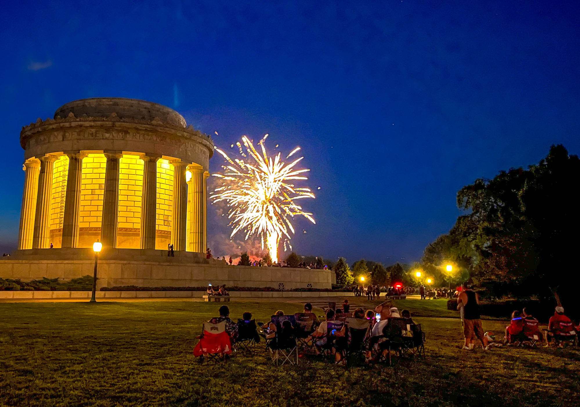 A crowd watching nighttime fireworks over circular memorial building with prominent columns