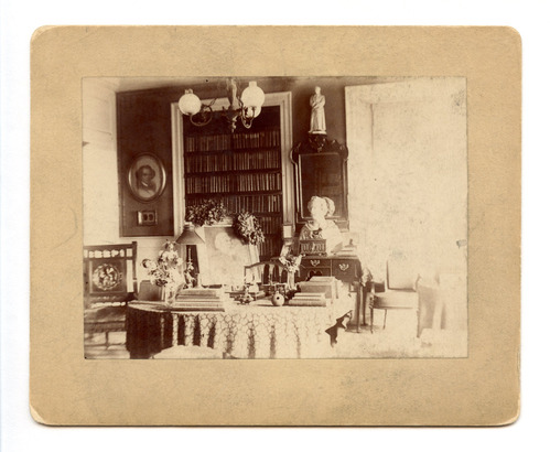 Black and white photograph of 19th century study including built in book case, and portrait on easel with wreaths.