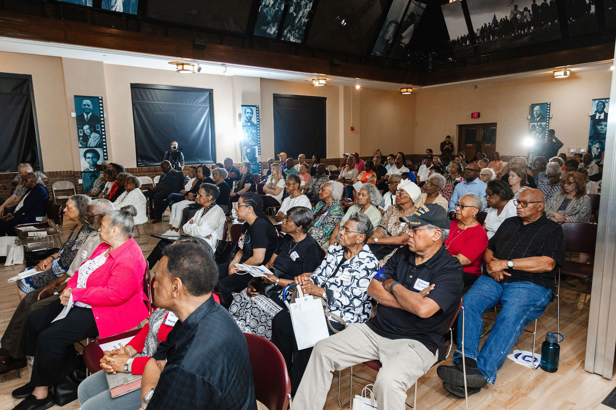 African American adults sit in maroon plastic chairs in a room with hardwood floors 
