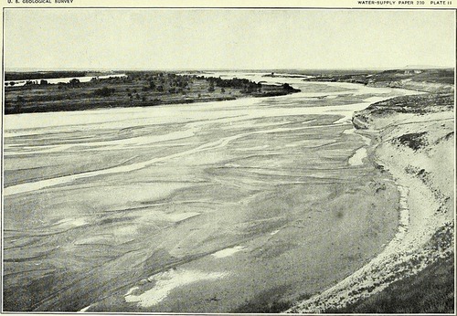 A broad, sand-bottomed river with an island of grass and scattered trees.