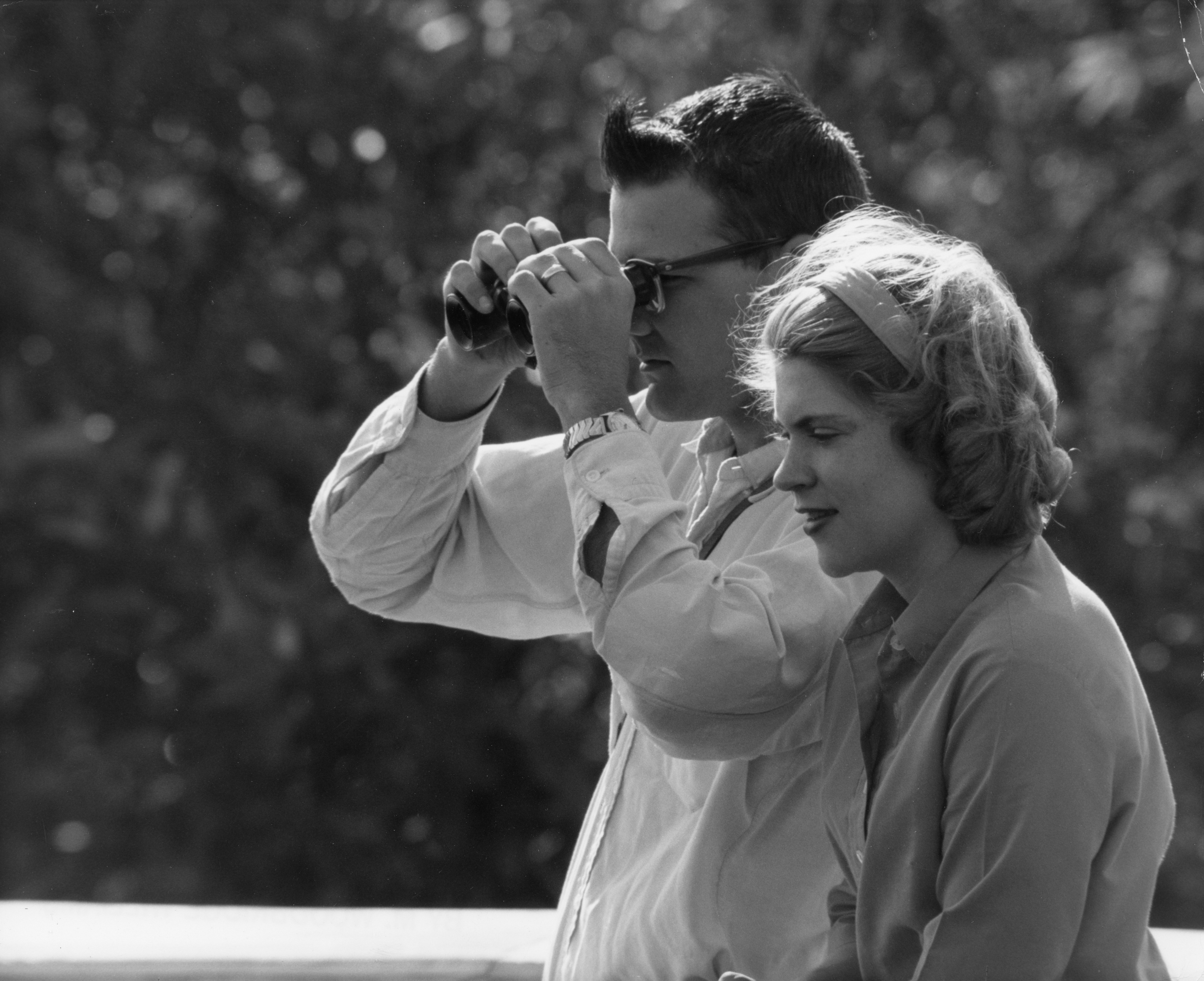 A close-up of a man and a woman in profile view, the man is looking through binoculars. 