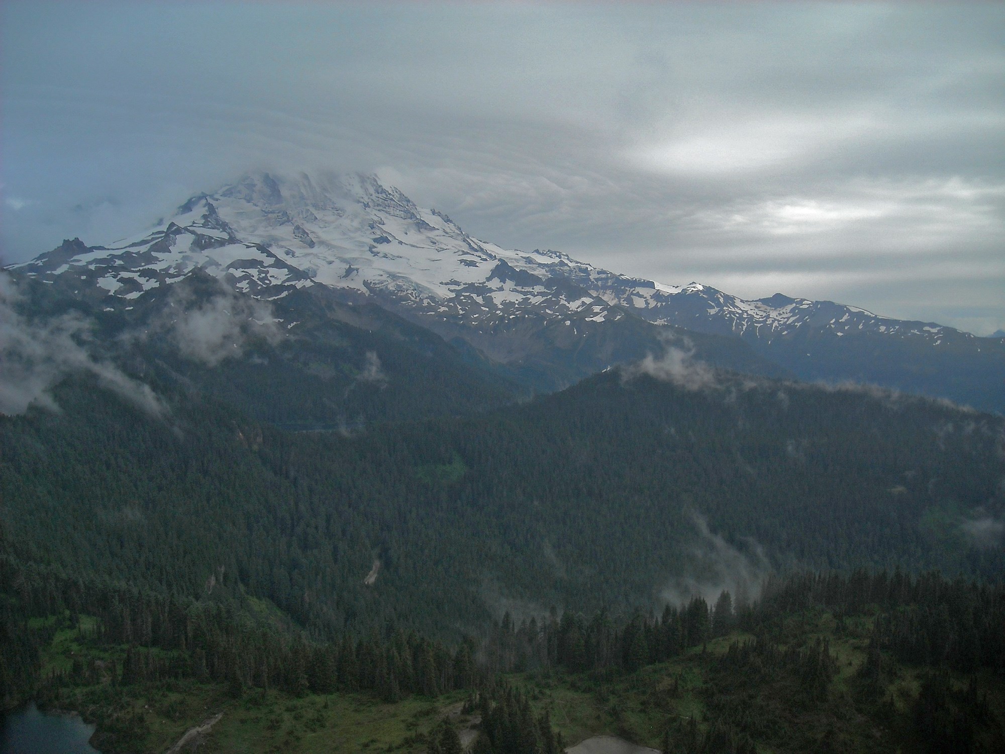 A tall, glaciated mountain peak wrapped in clouds above forested valleys and ridges. 