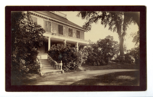 Black and white photograph of two story house with porch and shrubs.
