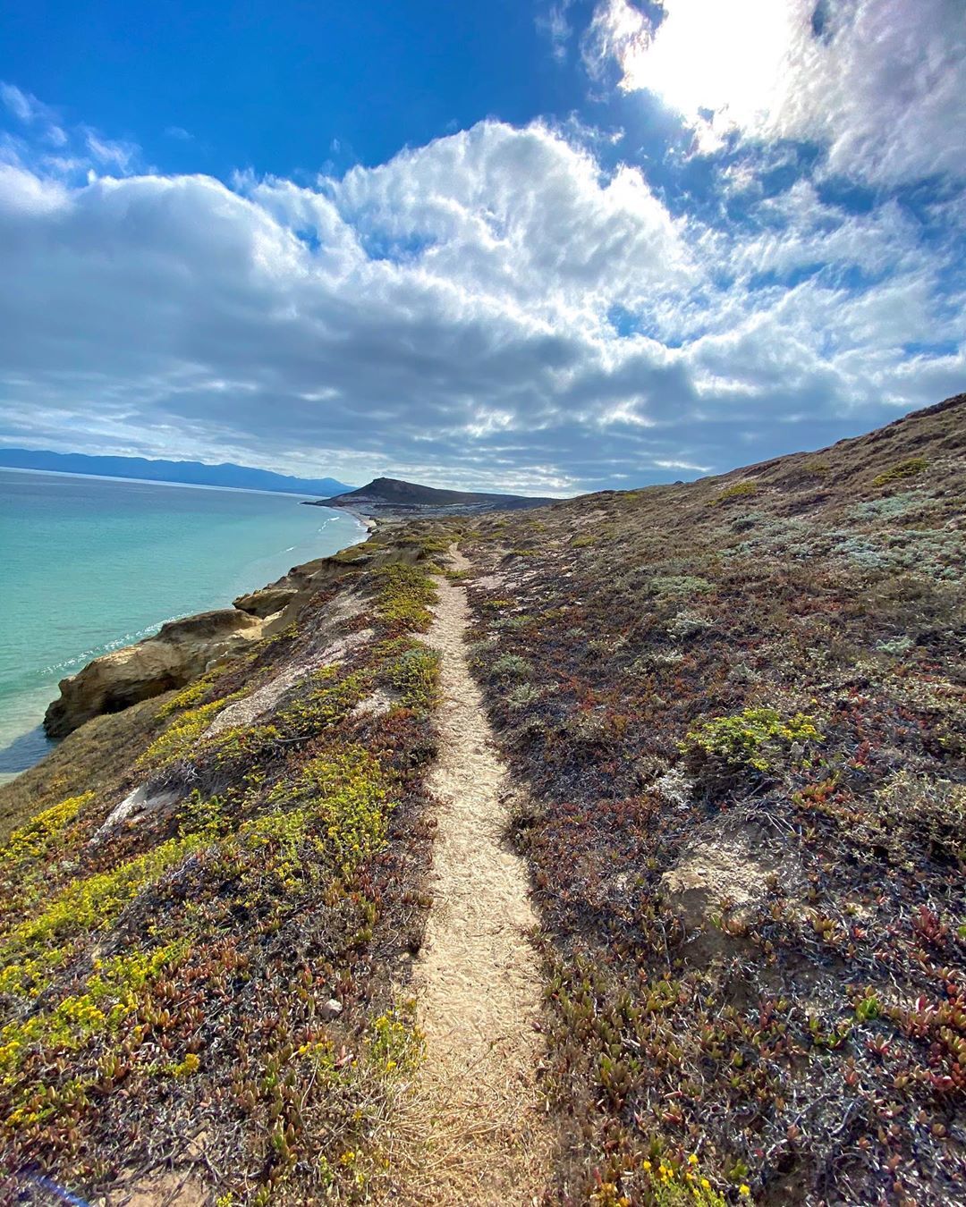 A narrow sandy trail cuts through the sloping hillside that meets crystal clear, calm ocean water. The hillside is covered in short, wiry brownish scrub with patches of light green and yellow vegetation. A bright blue sky and shiny sun are covered by long, billowy clouds