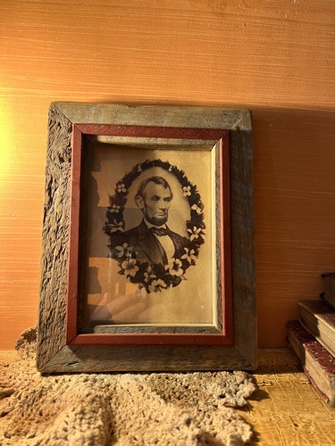 A small portrait of Abraham Lincoln in a wooden frame on a table leaning against a wall. 