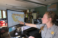 Alix points to a computer screen in her office. Alix is wearing a grey NPS collared shirt. She is pointing to the Maxtent Model she has recently run on her computer. The image on the computer screen shows yellow splotches on top of a map of Great Smoky Mountain National Park.