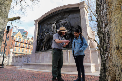 Park Ranger shows visitor how to connect to the National Park Service App. The pair stands facing the camera looking at a cell phone. They are in between two large trees and in front of a large Civil War Memorial.