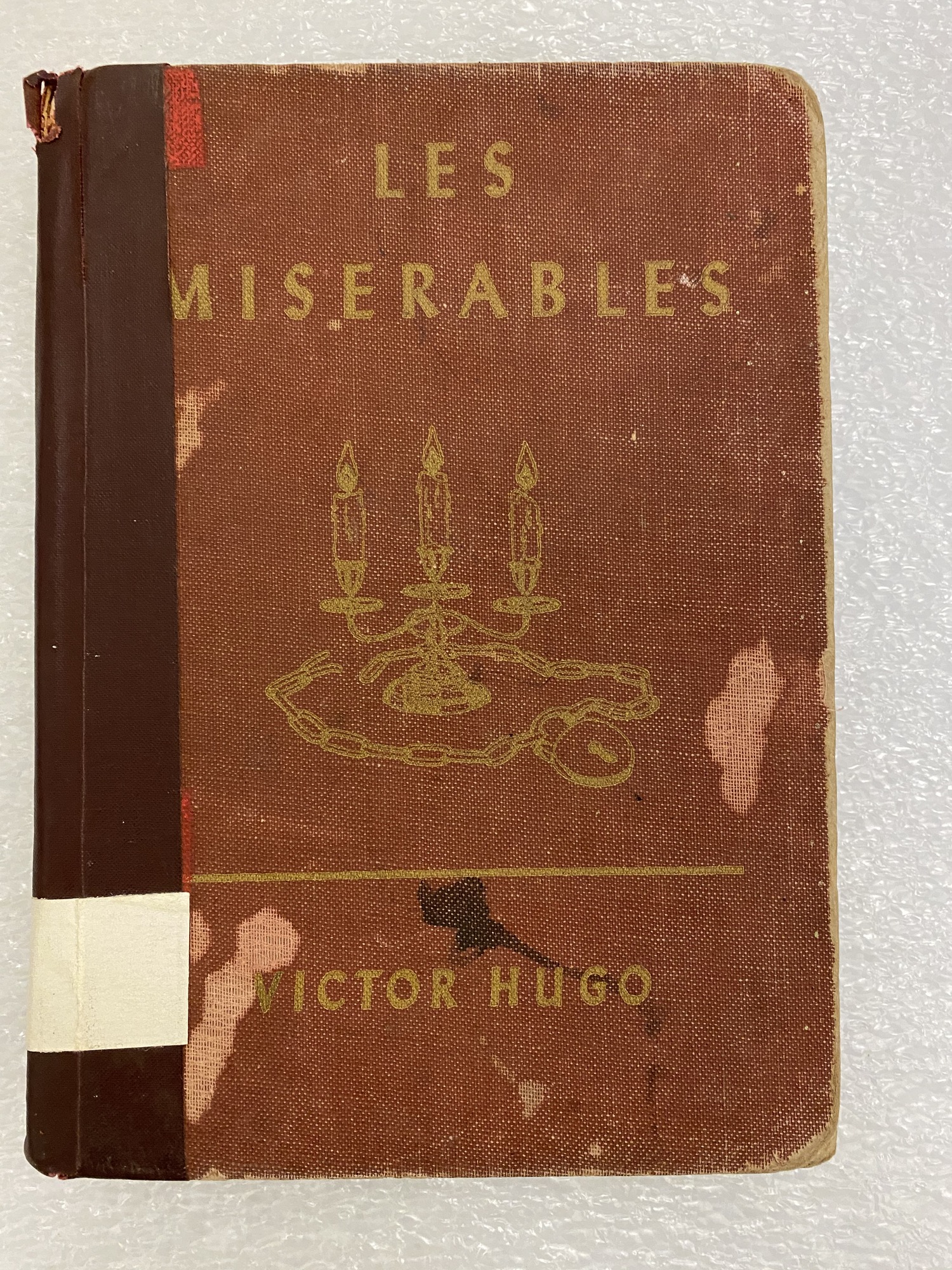 Cover of the book Les Miserables. The cover is a faded red with gold lettering and an image of a candle stick holder with a chain and lock at its base. The spine side of the book has been repaired with dark red book tape