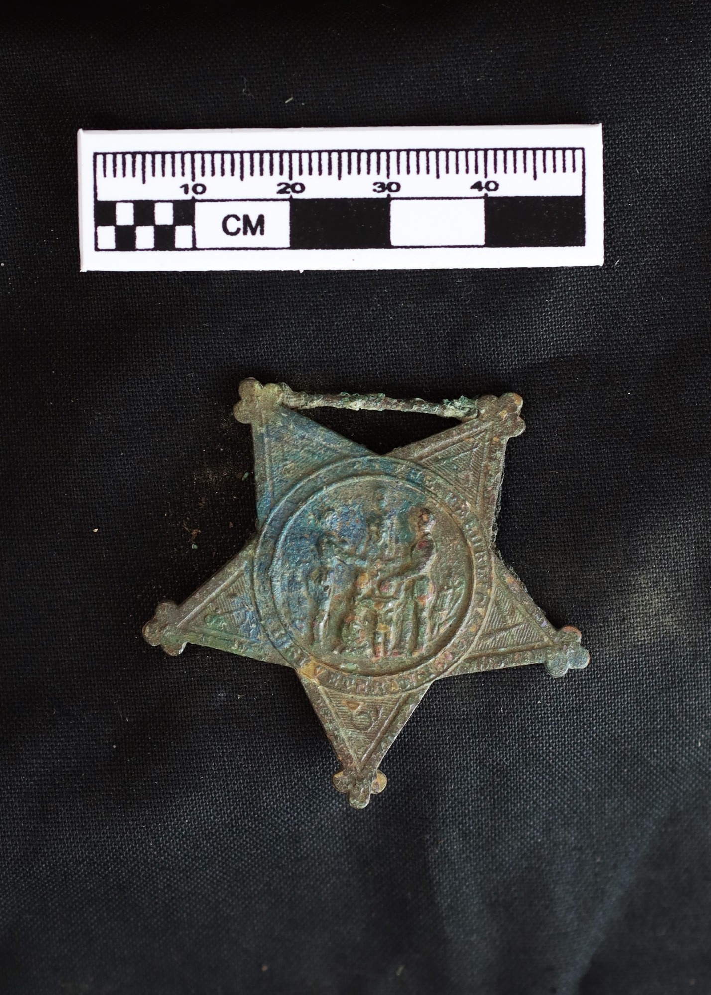 5-point star shaped badge with a greenish colored patina. Three figures in center of star. On black background with ruler showing it as 4-5 cm in size