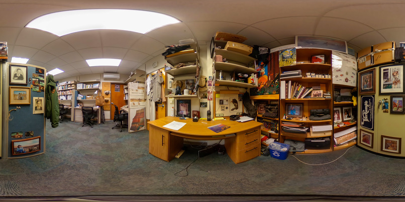 360 photo of an interpretive ranger's desk and workspace. Above the desk are book shelves. A second set of bookshelves fills the wall on the immediate right of the desk. There are books, an antique typewriter, rocks, figurines, and a variety of other nick-nacks filling the shelves. Directly behind the desk is a partition that displays about a dozen photos of the ranger and his family, from different times during his life. 