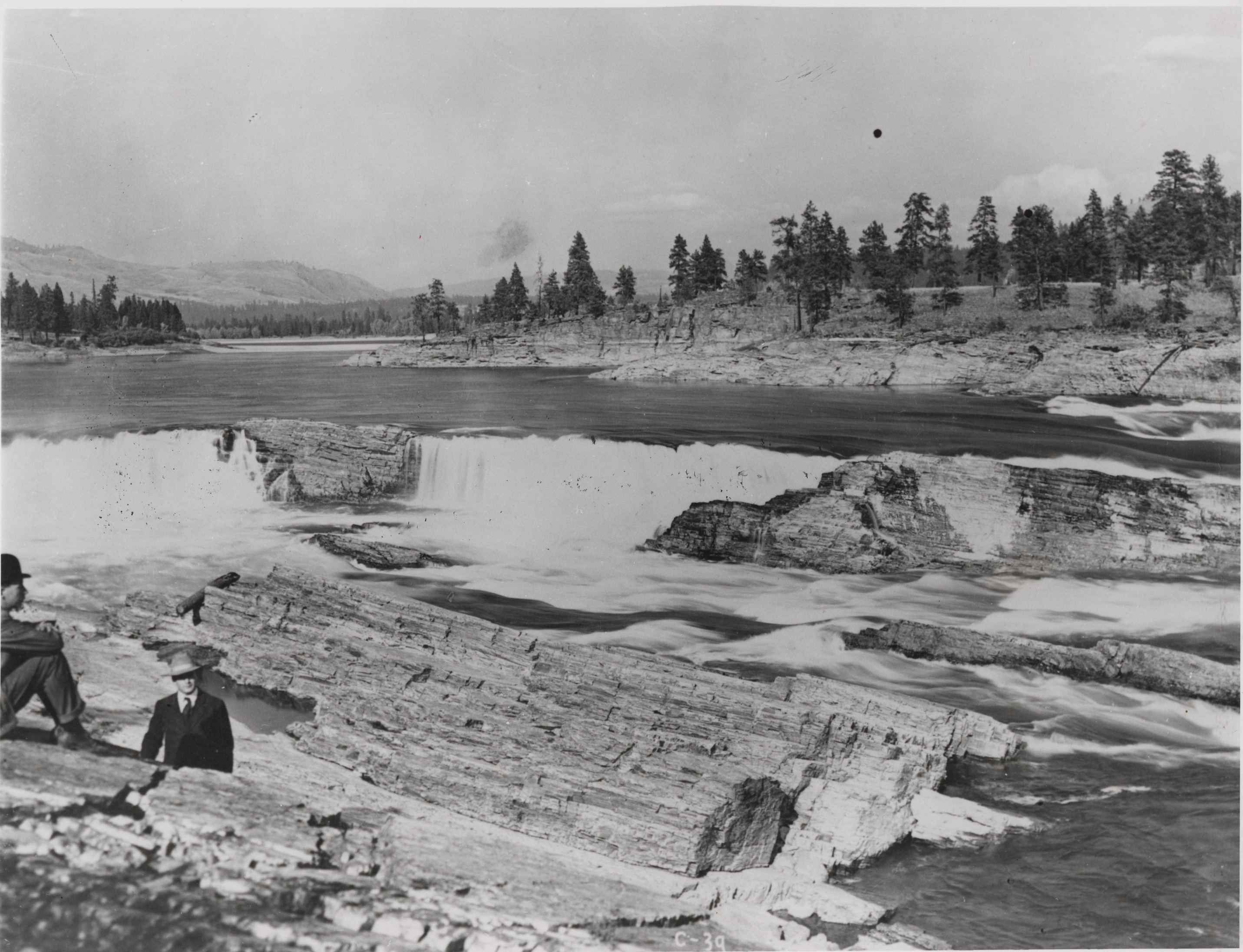 Black and white photograph of two men sitting on rocks next to a waterfall