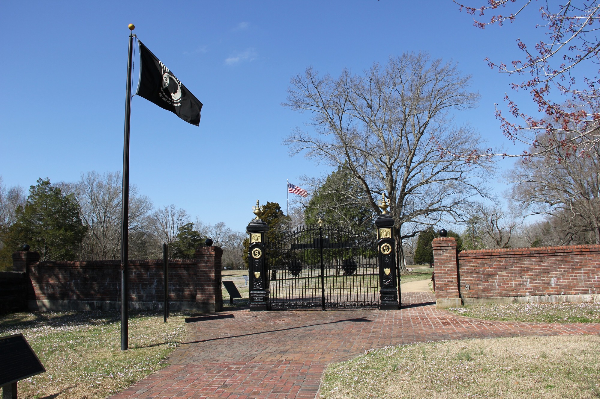 The main black iron gates with gold trim serve and brick wall mark the boundary of the national cemetery. A POW/MIA flag flies in the foreground.  