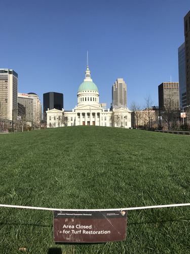 A domed courthouse stands beyond a slight dome of bright, neat grass, framed by modern city high rises and a clear blue sky. 