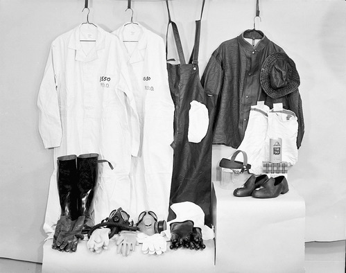 Black and white photo of coats and an overall hanging against a wall. In front are multiple pair of gloves, face masks, and shoes. 