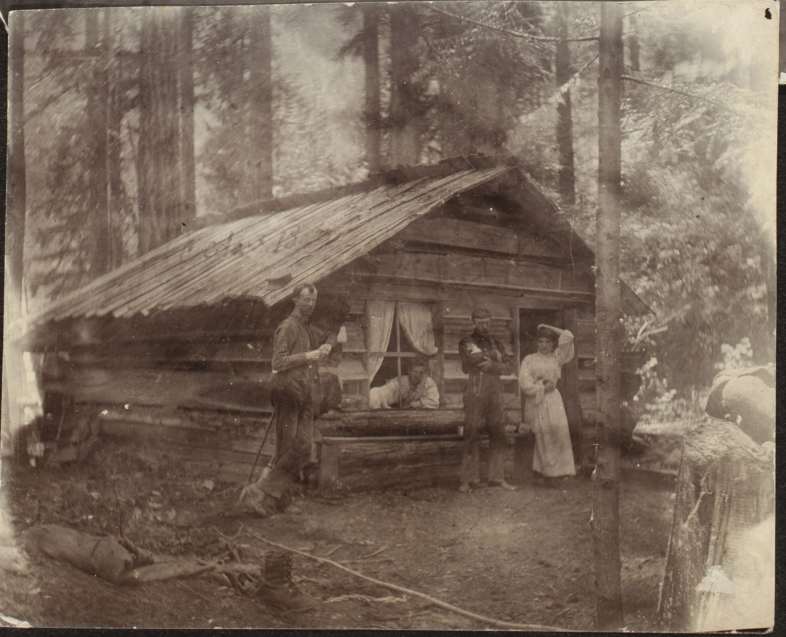 Two men and a woman standing outside a rough-hewn cabin in a forest. A third man pokes his head through a cabin window.