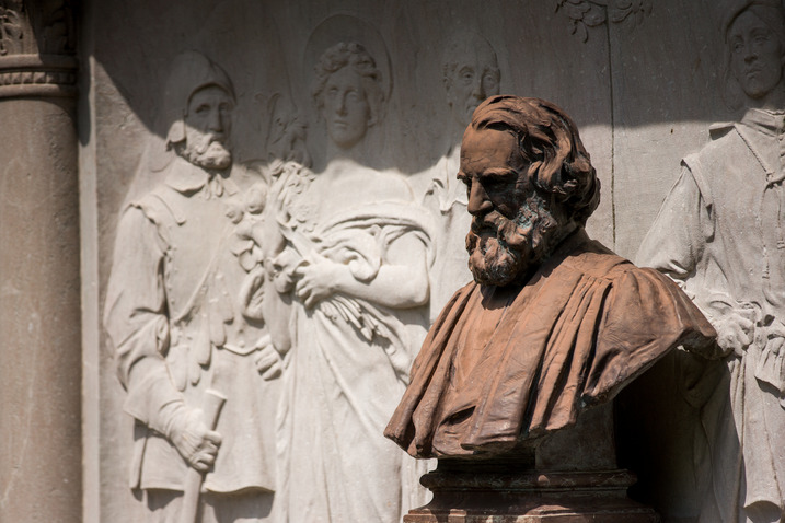 Bronze colored bust of Henry Longfellow with beard and academic robe, looking down. Figures carved in granite behind him include man in colonial clothing and angel carrying flowers.
