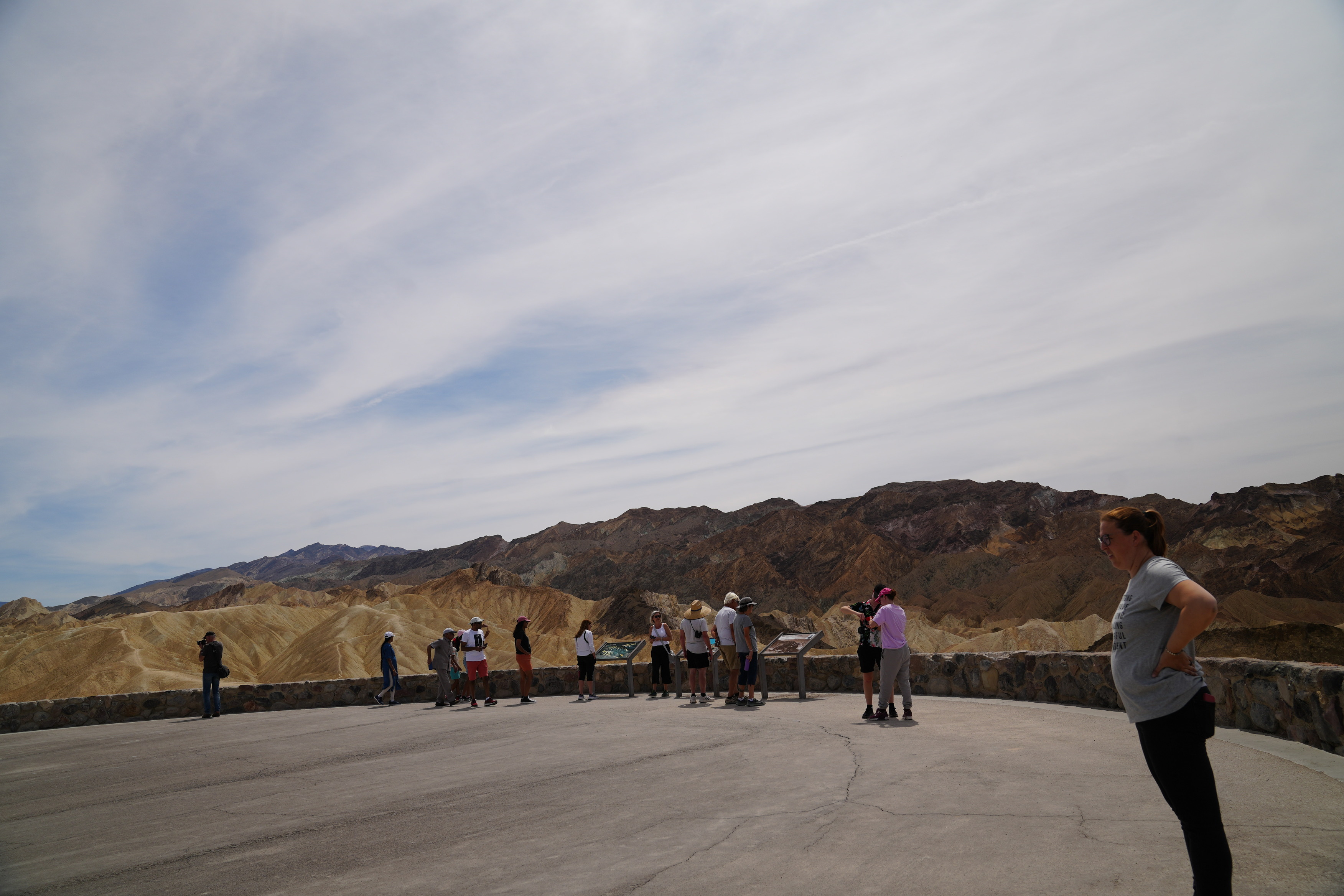 Zabriske Point viewpoint. The viewpoint is smooth asphalt at a slight angle going upwards. A thigh-height stone wall surrounds the viewing area. There are many waist-level informational signs. Yellow badlands and red geological formations can be seen. People are crowded around the edges looking at the view point.