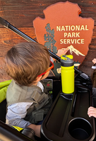 A child touches the mountain on a large arrowhead with the words, National Park Service. 