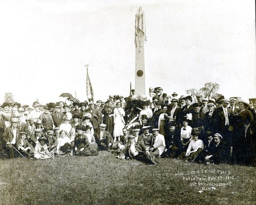 A black and white image of a group of people surrounding a tall stone monument. 