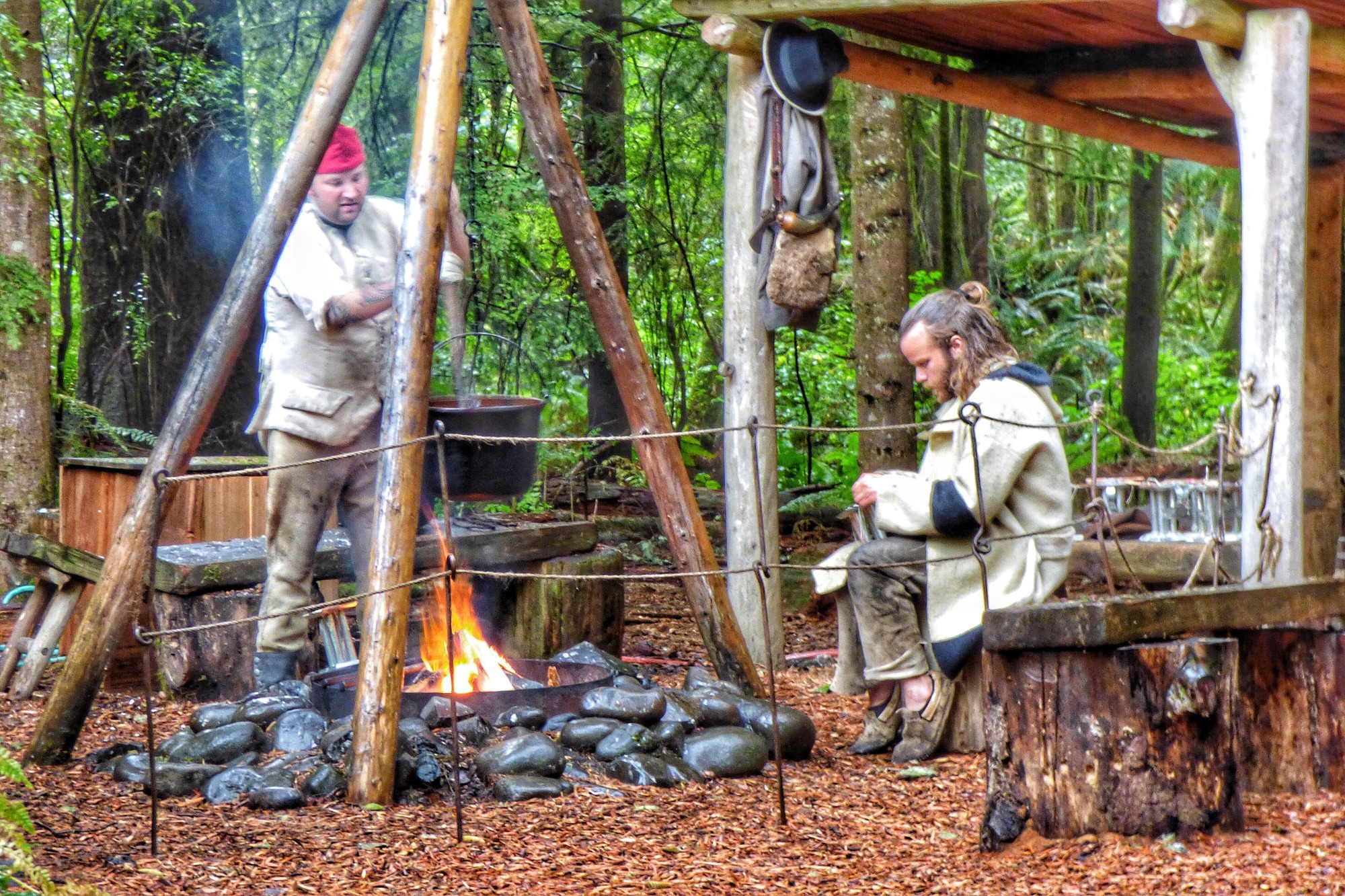 Two rangers in 1800s period attire sit around a campfire, rendering tallow in a pot hung over the fire.