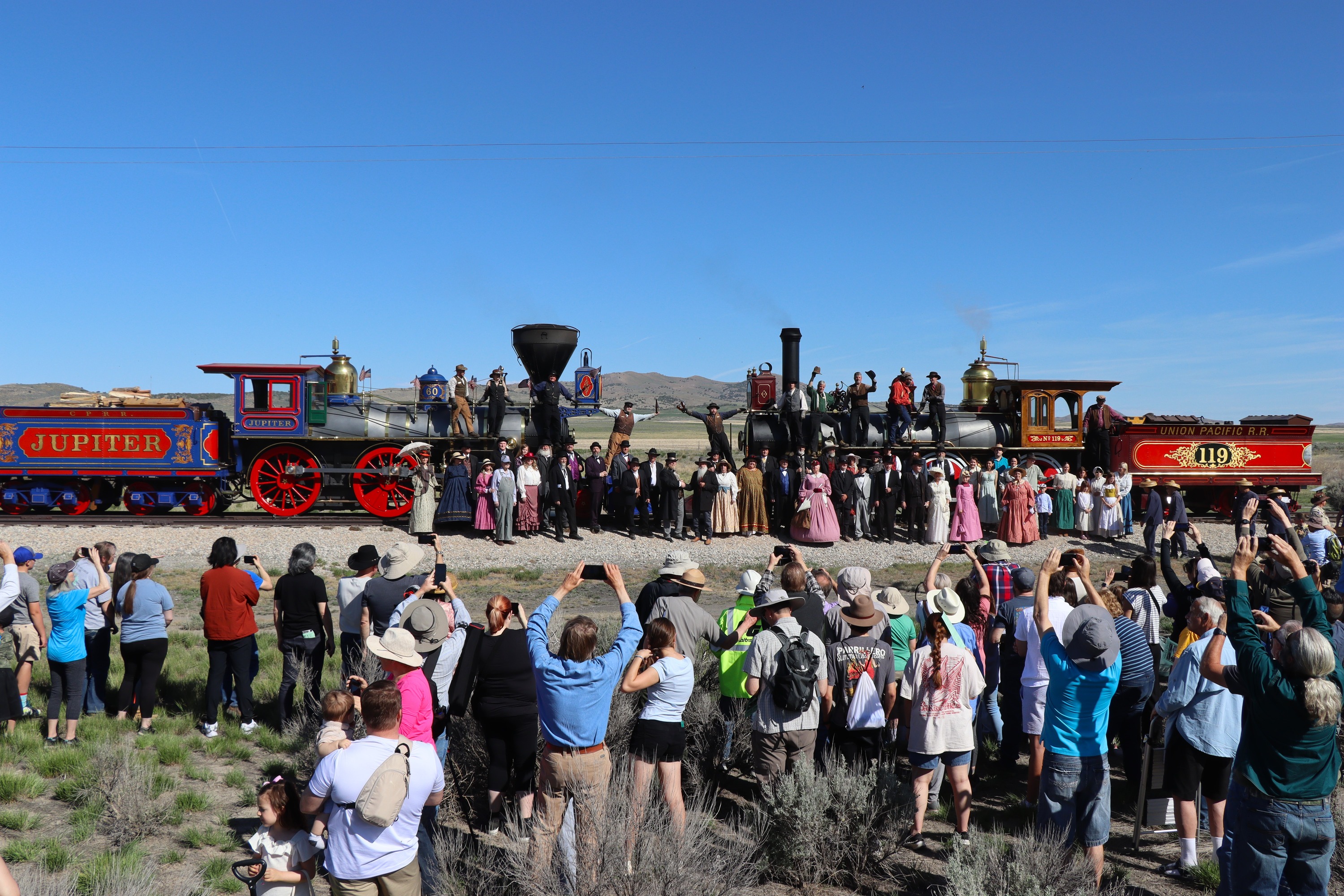 
Men and women in turn-of-the-century costumes stand in front of two vibrant historic locomotives, while two engineers raise champagne bottles. Visitors in foreground taking photos.