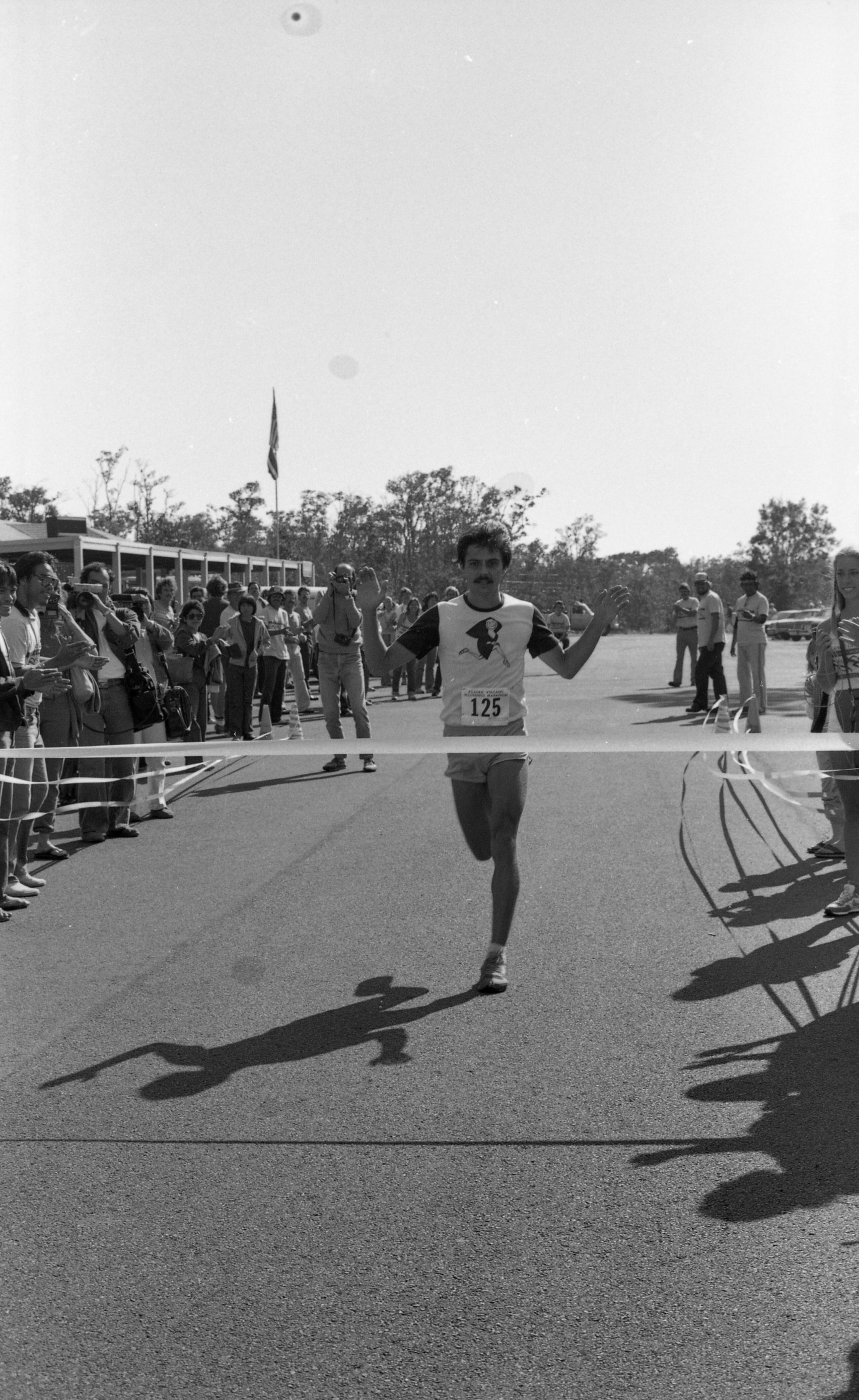 A black and white action shot of a man feet away from crossing the finish line during a race. The man is located in the center of the image. He is in mid run with his arms slightly bent at the elbow raised towards his shoulders. The man is wearing a t-shirt with dark colored sleeves and a light-colored body. There is a smiley face graphic wearing a cape printed on the front of his shirt. Affixed to his shirt is a race bib. The bib reads, "Kīlauea Summit Wilderness Marathon 125." He is also wearing running shorts and sneakers and has a moustache. In front of him running the length of the photo is the finish line ribbon. The finish line is located in the front parking lot of the Kīlauea Visitor Center, which can be seen behind a crowd lined up across its front lanai. On the right side of the image there are more people gathered cheering the runner on.