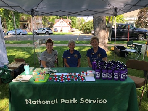 three ladies sit behind a table with a green table cloth and art supplies at an outdoor park