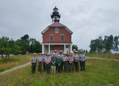 A group of people wearing gray shirts and green pants pose in front of a lighthouse and brick keeper's quarters. 