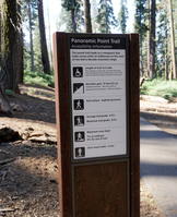 A metal panel with text and pictographs has information regarding accessibility for visitors with disabilities. To the side of the metal sign is a paved walking path that cuts through trees and low growing bushes. 