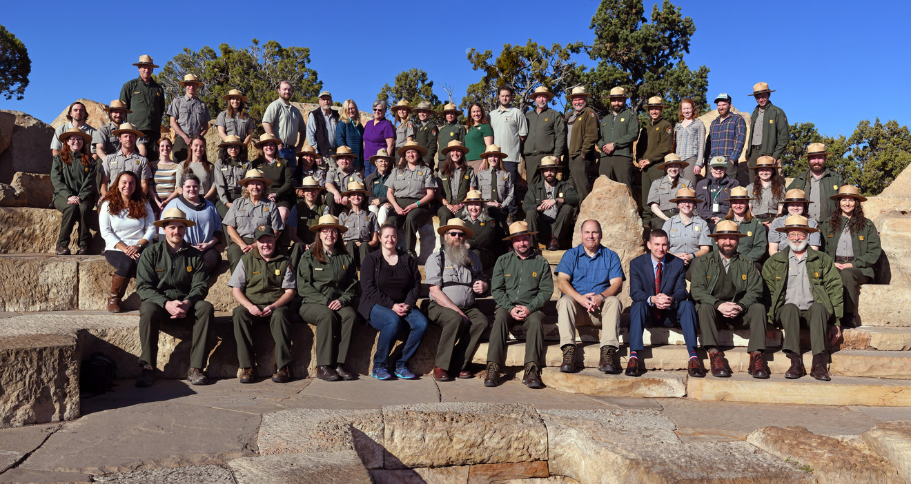 A group photo of around 45 individuals in four rows, most wearing NPS uniforms and sitting on the steps of a stone amphitheater
