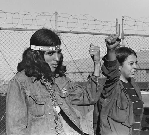 A male occupier wearing denim jacket, beaded headband, and feathered fetish stands next to female occupier, both holding their fists high in front of barbed-wire topped fence at a pier in san Francisco 