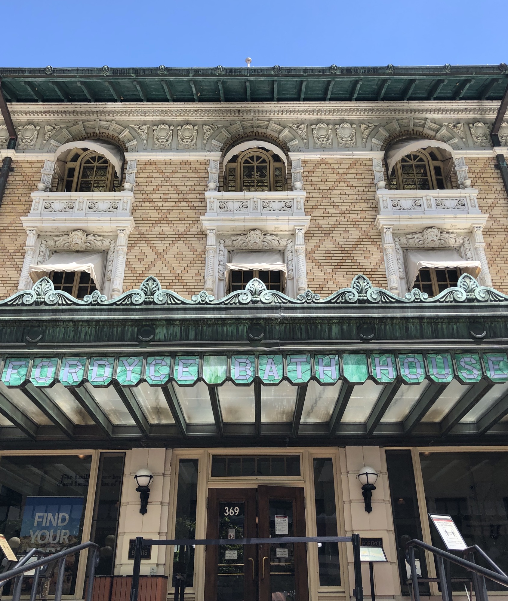 Large 3 story building with ornately carved windows and a large green porch with a stained glass panels that spell out "Fordyce Bath House."