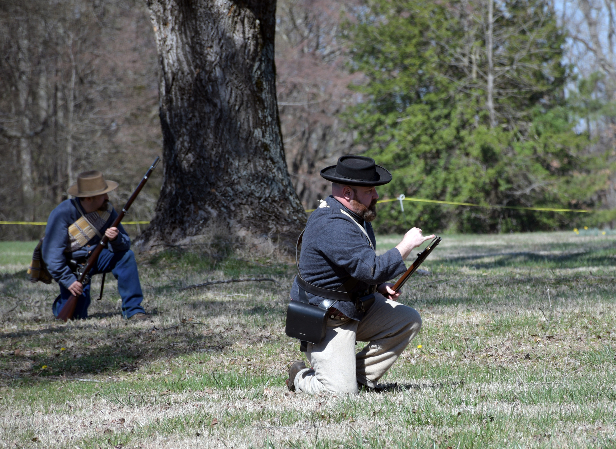 Two men wearing Confederate uniforms, both kneeling, the man in the foreground uses his pinky finger to the push a ramrod back into place while the second made in the background watches.