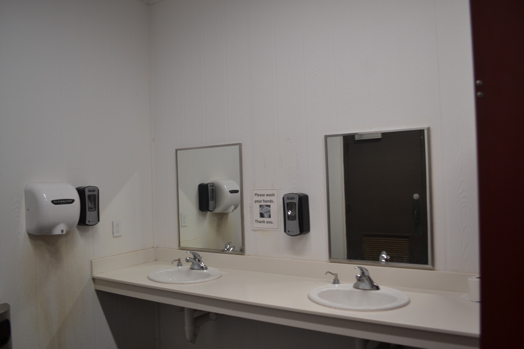 An all-white room with sinks on the left side and stalls to the right. A white countertop with two sinks and two mirrors each. To the left of the sinks is a soap dispenser, hand dryer and trashcan along this wall. 