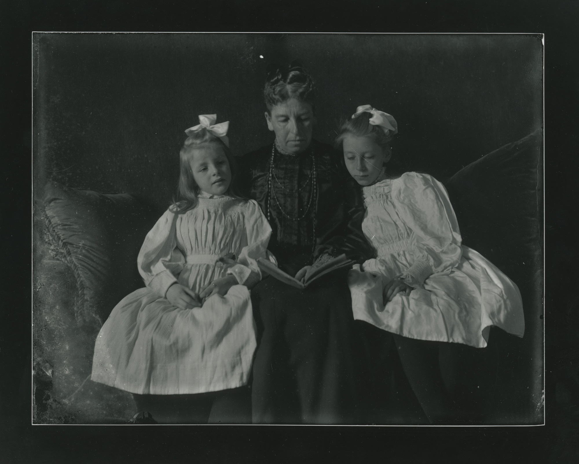 A white woman sits and reads on a couch in between two young girls.